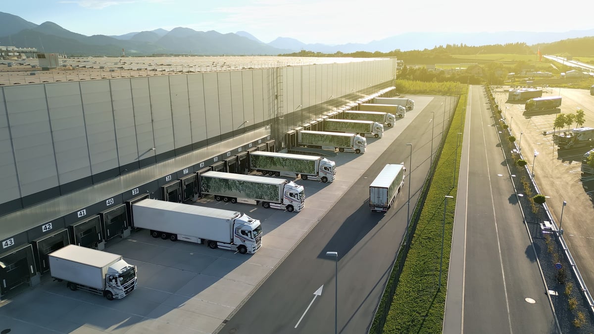 An aerial shot of aerial view of a bustling distribution warehouse with multiple trucks lined up at loading docks, set against a scenic mountain backdrop. The scene highlights efficient freight transportation and logistics operations, emphasizing modern infrastructure and cargo management.