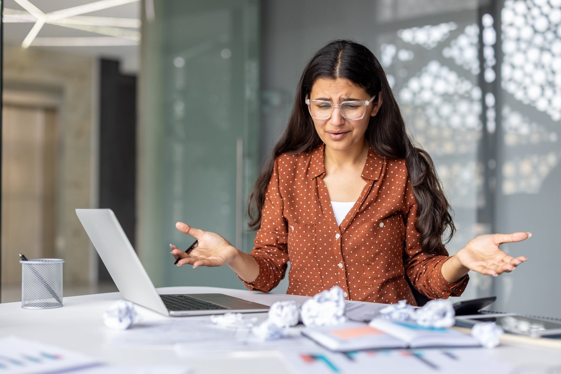 Stressed businesswoman gesturing in frustration while looking at her laptop, surrounded by crumpled papers on her desk