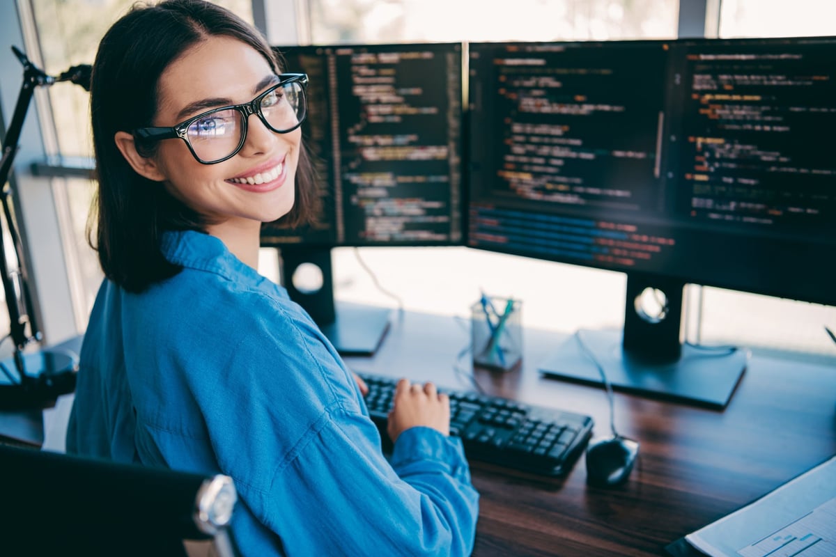 A focused young professional works at a desk with multiple monitors displaying code, keyboard and mouse in a bright loft office environment