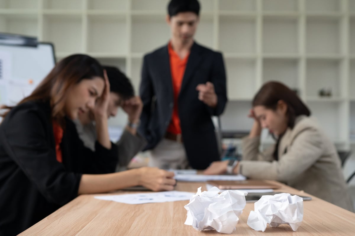 Stressed employees holding their heads in frustration during a tense workplace meeting with a standing manager