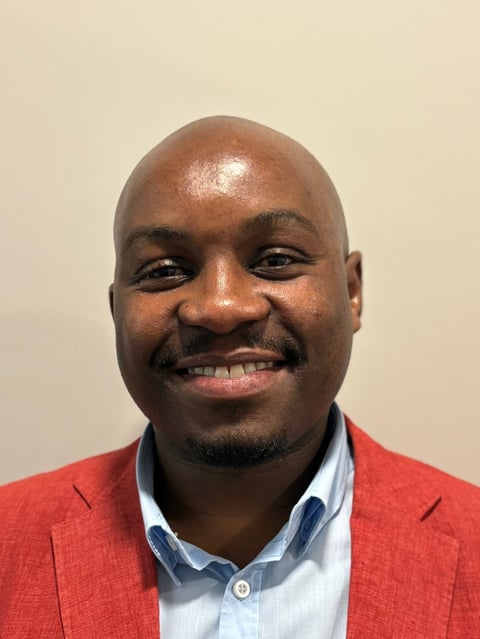 Smiling man with bald head wearing a red blazer and light blue button-up shirt against a beige background