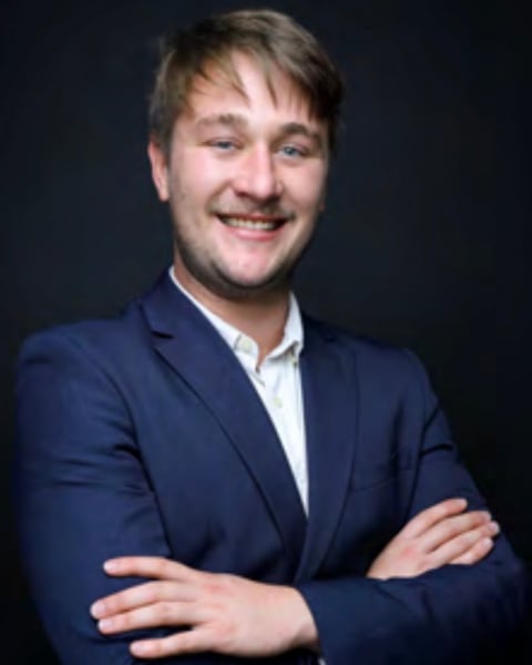 Professional headshot of a smiling man with light brown hair wearing a dark blue blazer and white shirt, arms crossed against a dark background
