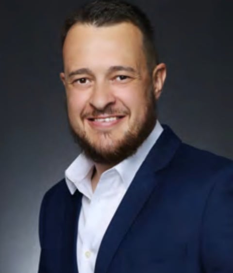 Professional headshot of a smiling man with a beard wearing a dark blazer and white shirt against a gray background