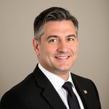 Professional headshot of a smiling man in a black suit and dark tie against a neutral beige background