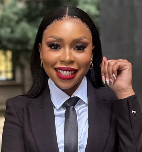 Professional woman in black blazer and blue shirt with tie, wearing bold red lipstick and silver hoop earrings, smiling at camera