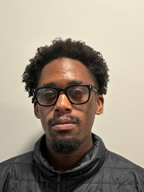 Headshot of a man with dark curly hair, glasses, and a black puffy jacket against a neutral beige background