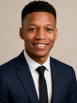 Professional headshot of a smiling man wearing a navy blazer, white dress shirt, and black tie against a neutral background