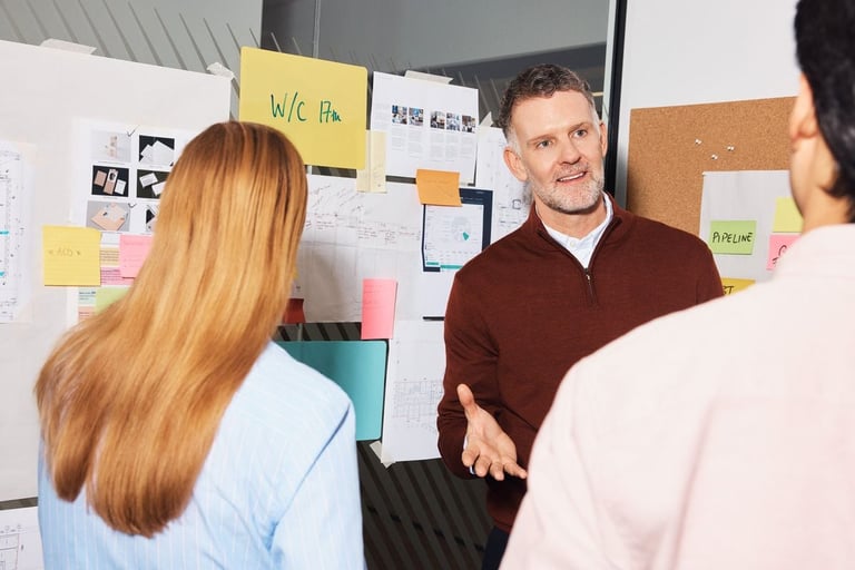 Team members gathered around a whiteboard with sticky notes during a collaborative brainstorming or planning meeting in an office setting