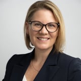 Professional headshot of a woman with shoulder-length blonde hair, glasses, and a dark blazer smiling at the camera