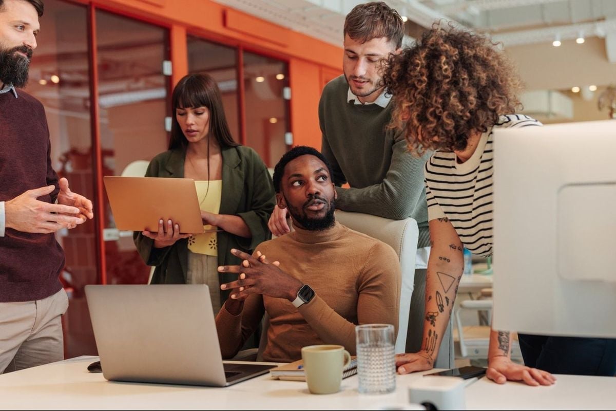 Diverse team of five professionals collaborating in an office setting, gathered around a laptop during a business discussion or presentation.